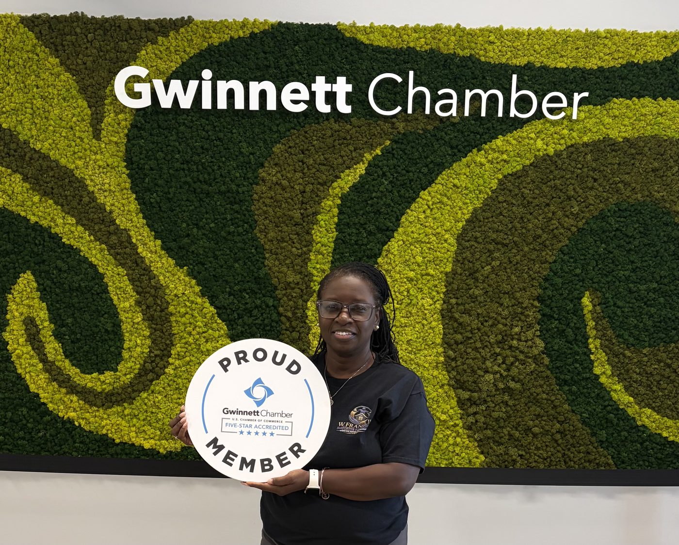 Wander Franklin smiling and holding a round Gwinnett Chamber Proud Member, Five-Star Accredited sign in front of the Chamber's moss wall display.