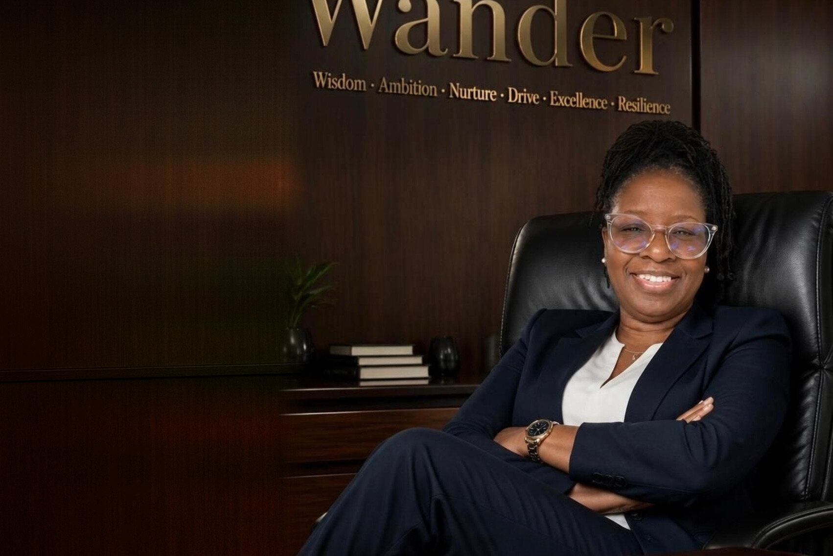 Wander Franklin smiling in a navy suit, arms crossed, seated in a black leather chair in her wood-paneled office under the gold lettering Wander.
